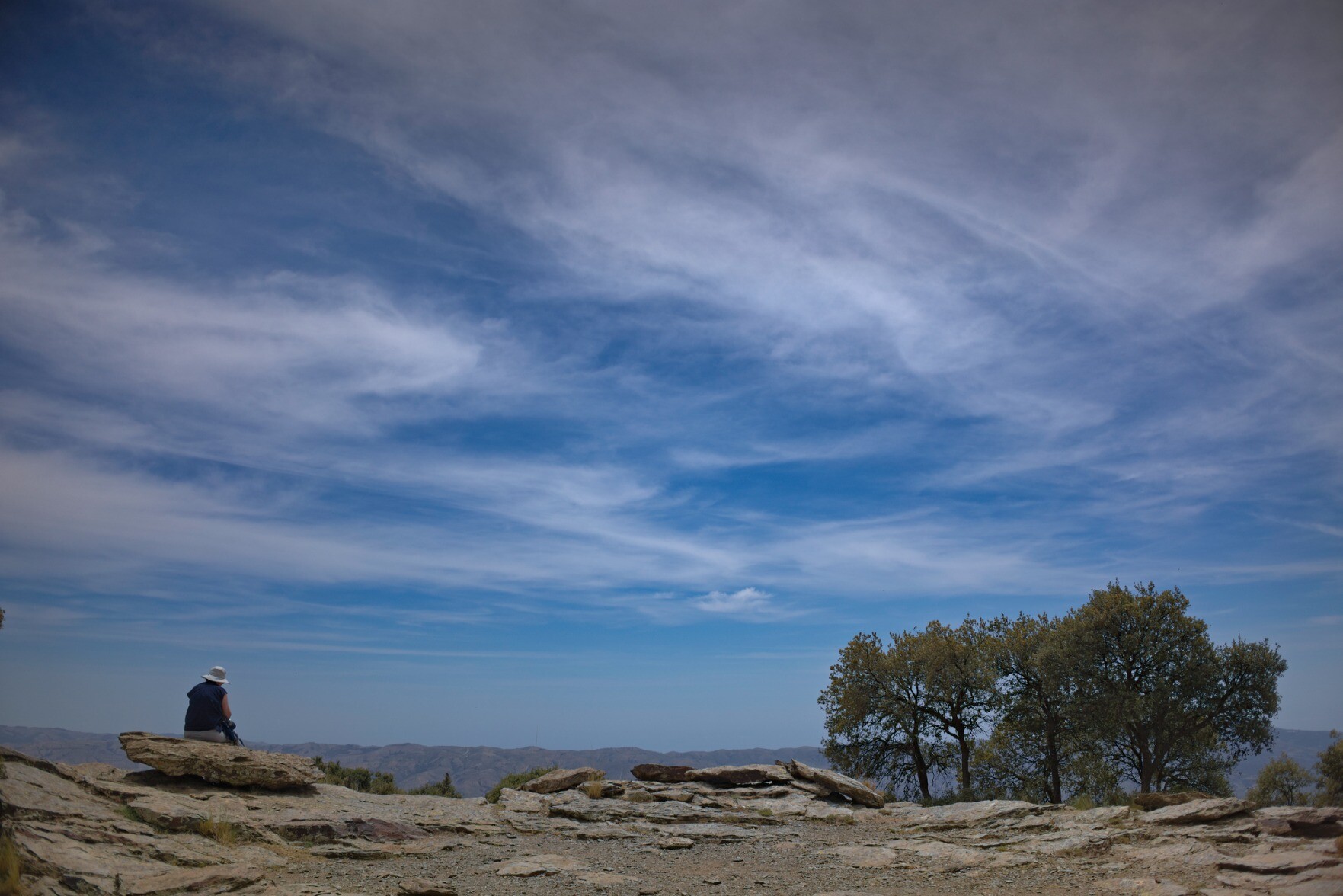 A person sits at the bottom left of the image enjoying the view. Above is a huge sky filled with wispy clouds. Some trees are to the right