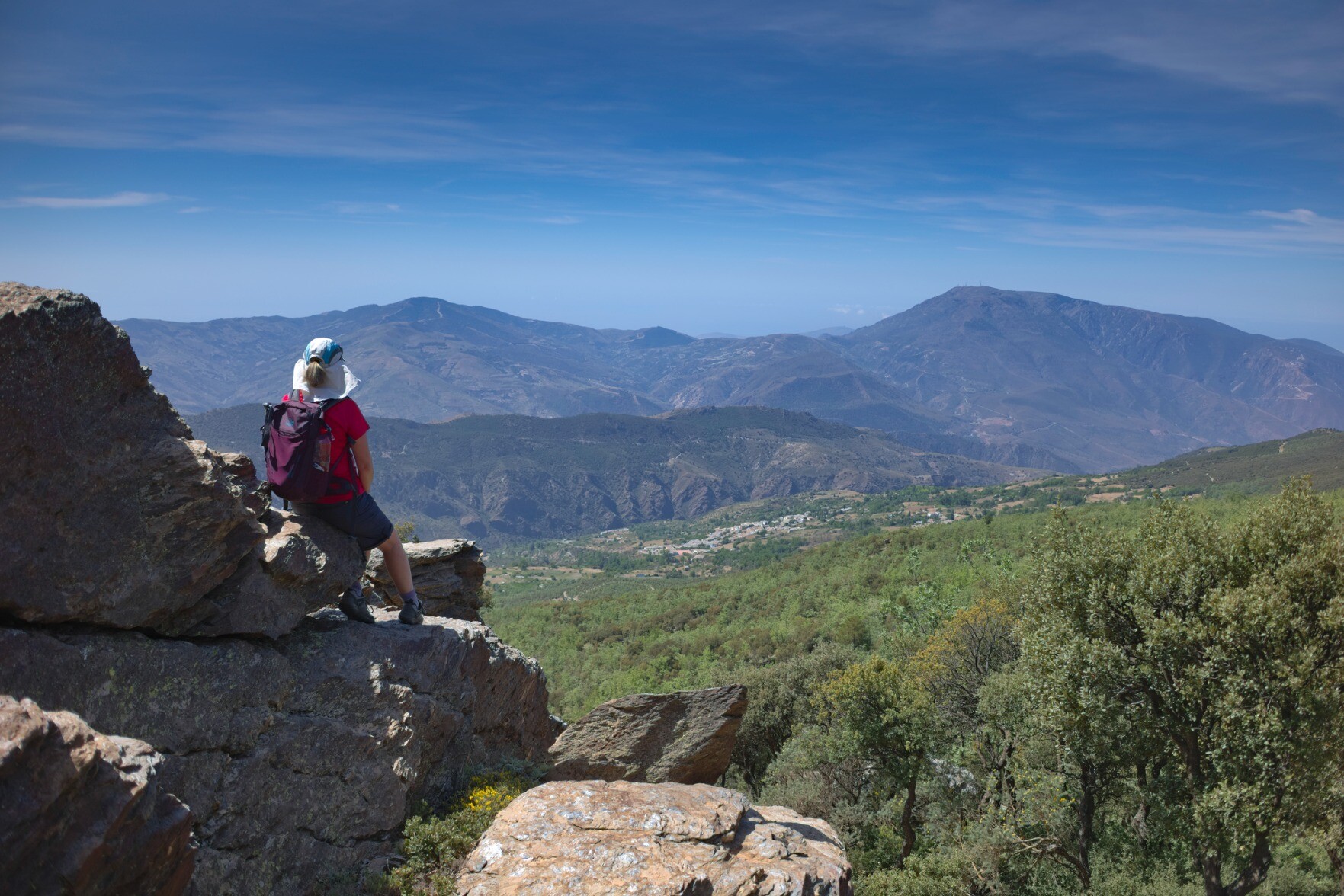 A person sits on a rocky outcrop looking at the view of forests, mountains and blue sky