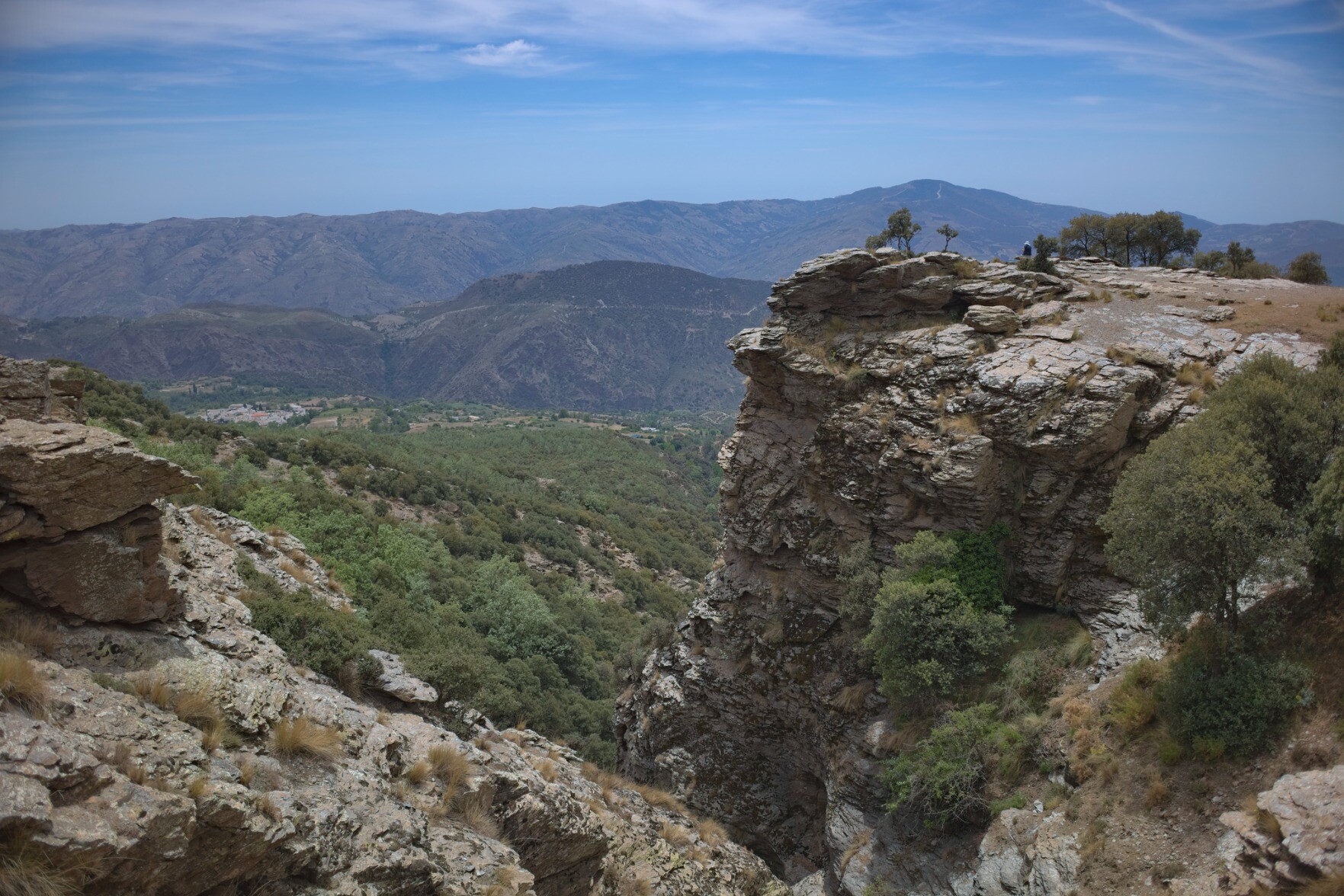 In the foreground is a rocky gorge, the right hand rocks have trees on top. In the distance are green forests and a range of low mountains