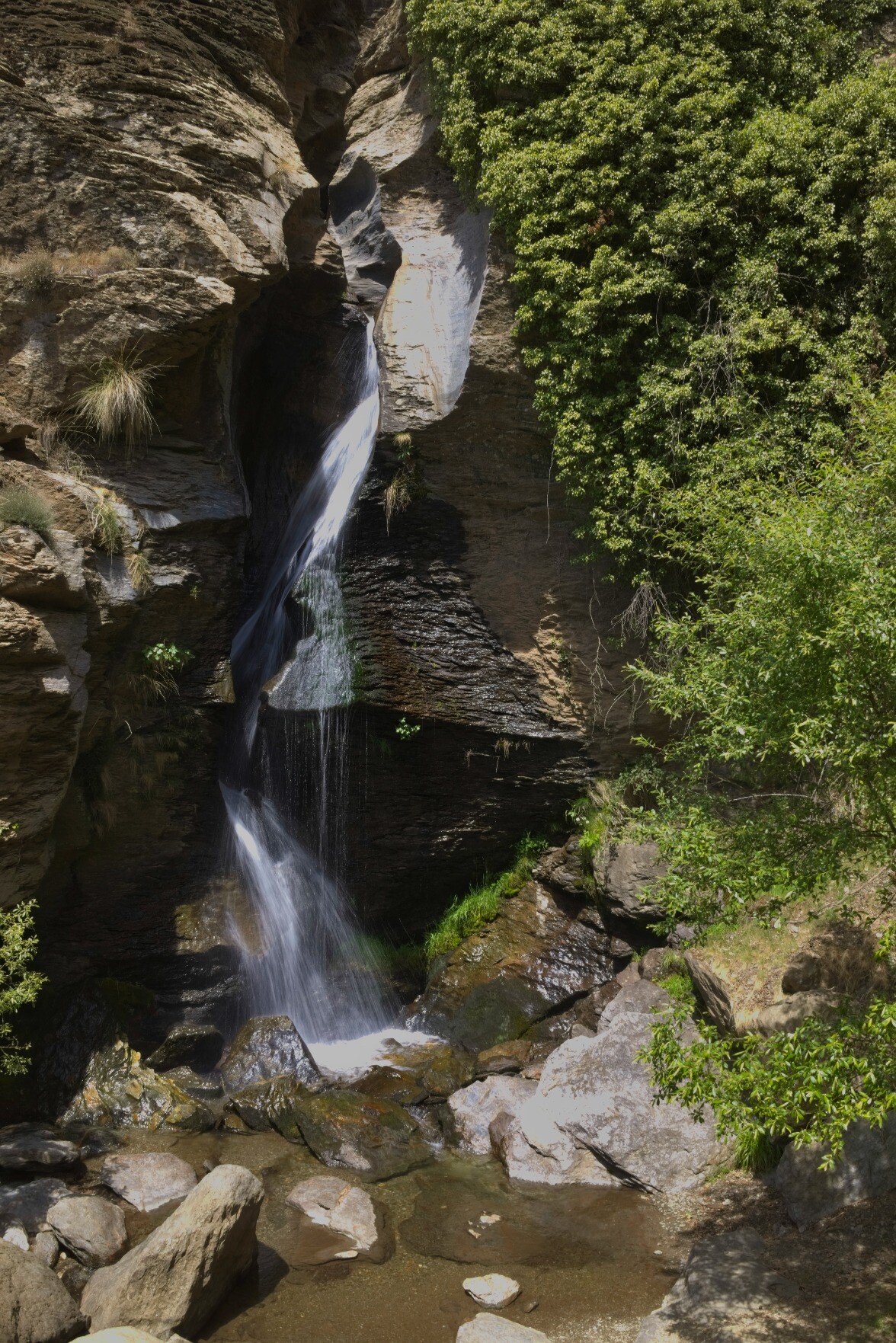 Water plunges down through a narrow gorge to form a waterfall. To the right is a lush green area of vegetation