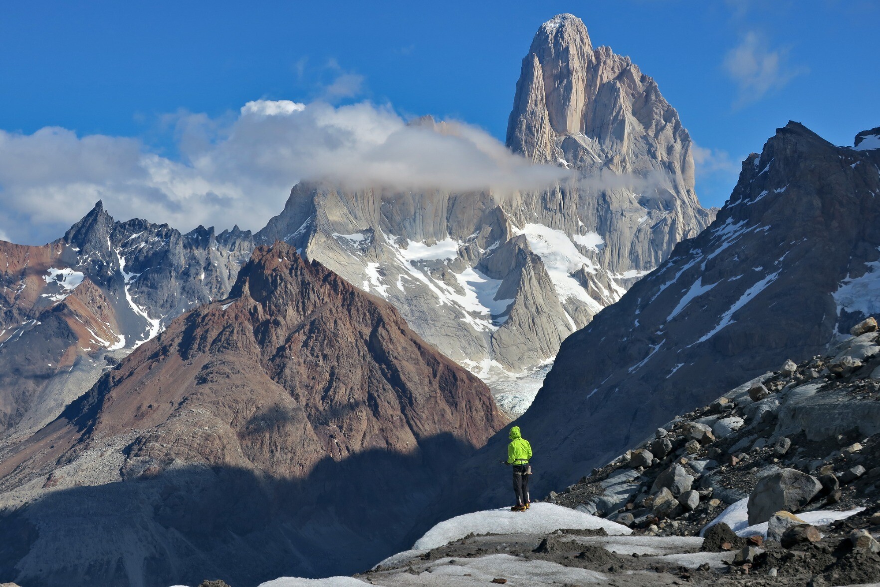 A man with green jacket is stood on a small rise below the giant mountain of Mt Fitzroy in Patagonia. Blue skies all around