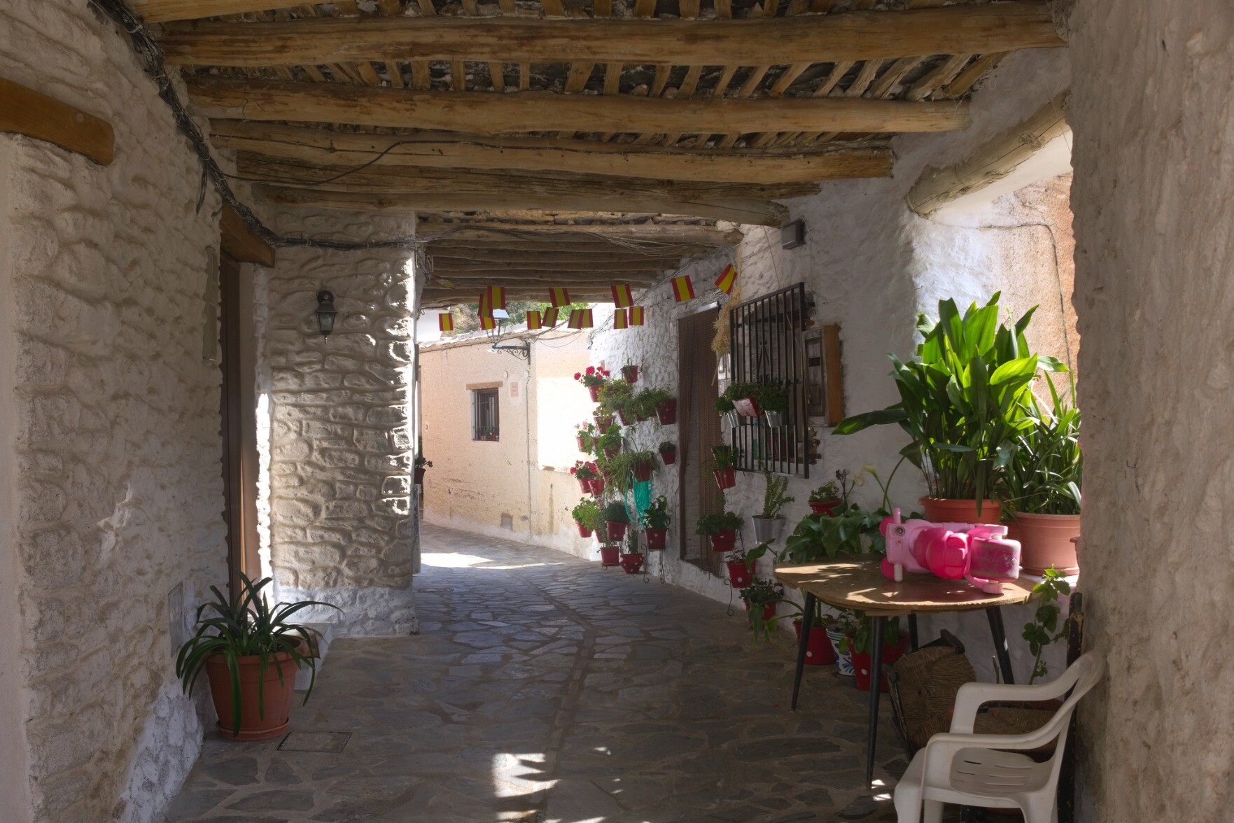 A narrow alleyway with old an building on the left and some open windows on the right. Flowers and colors adorn the sunlit building on the far right