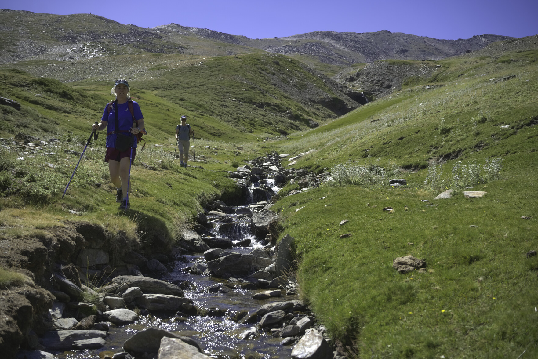 Two hikers walk down a beautiful green valley with a stream running down beside them