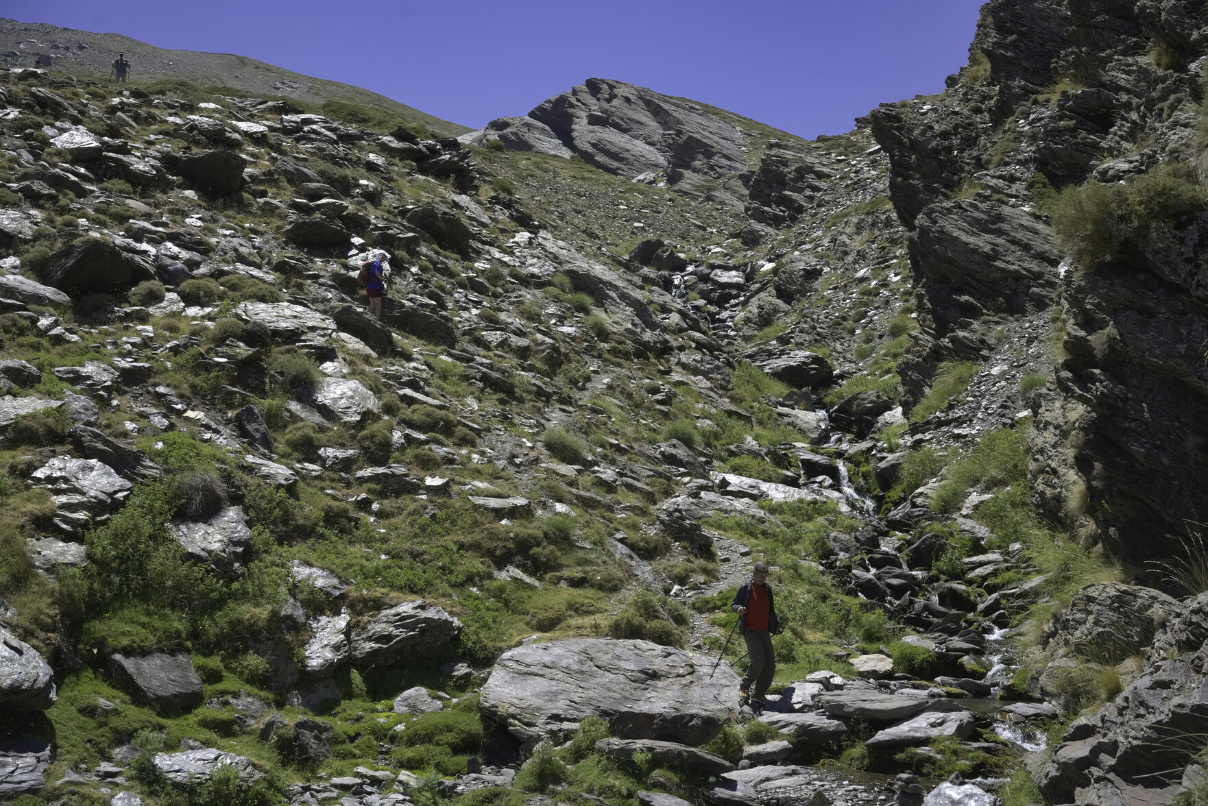 People descending a rocky rough valley with a small stream and a large cliff to the right