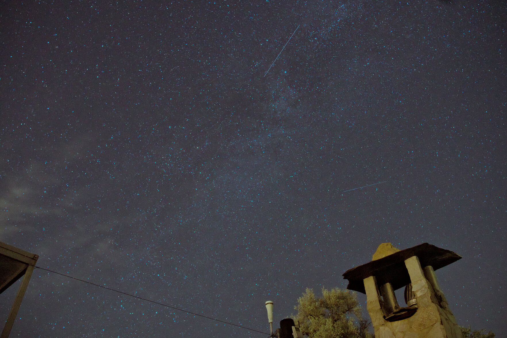 A night sky full of stars with a few satellite lines showing. At the foot of the image is a chimney and a weather station