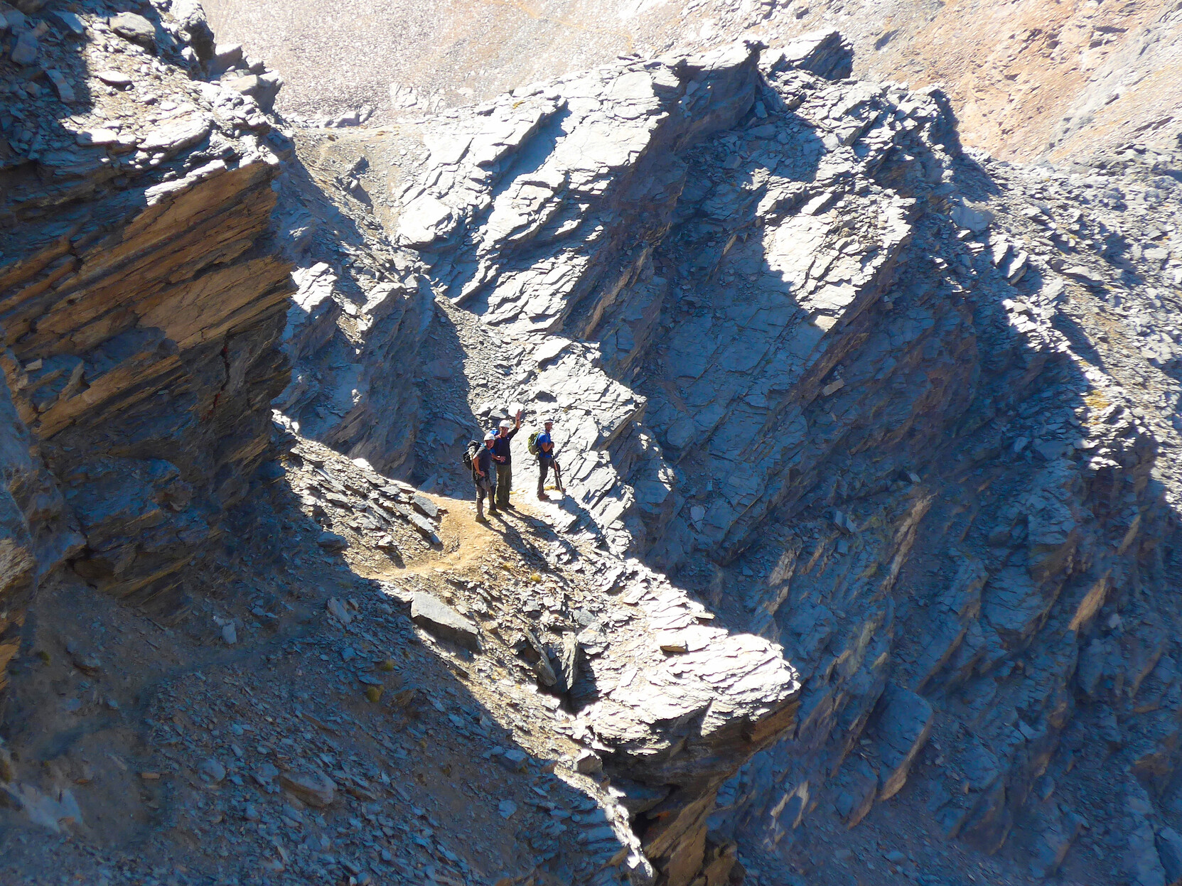 Some hikers stand on a narrow ledge illuminated in the sunlight. Steep cliffs are above them and below them.