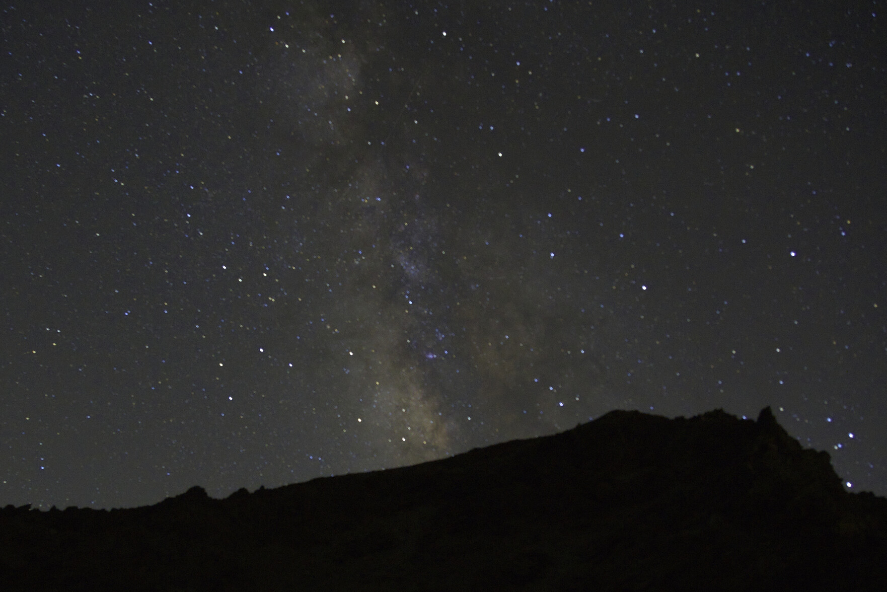 A dark mountain peak is in the lower photo. Above the Milky Way and hundreds of stars illuminate the skies