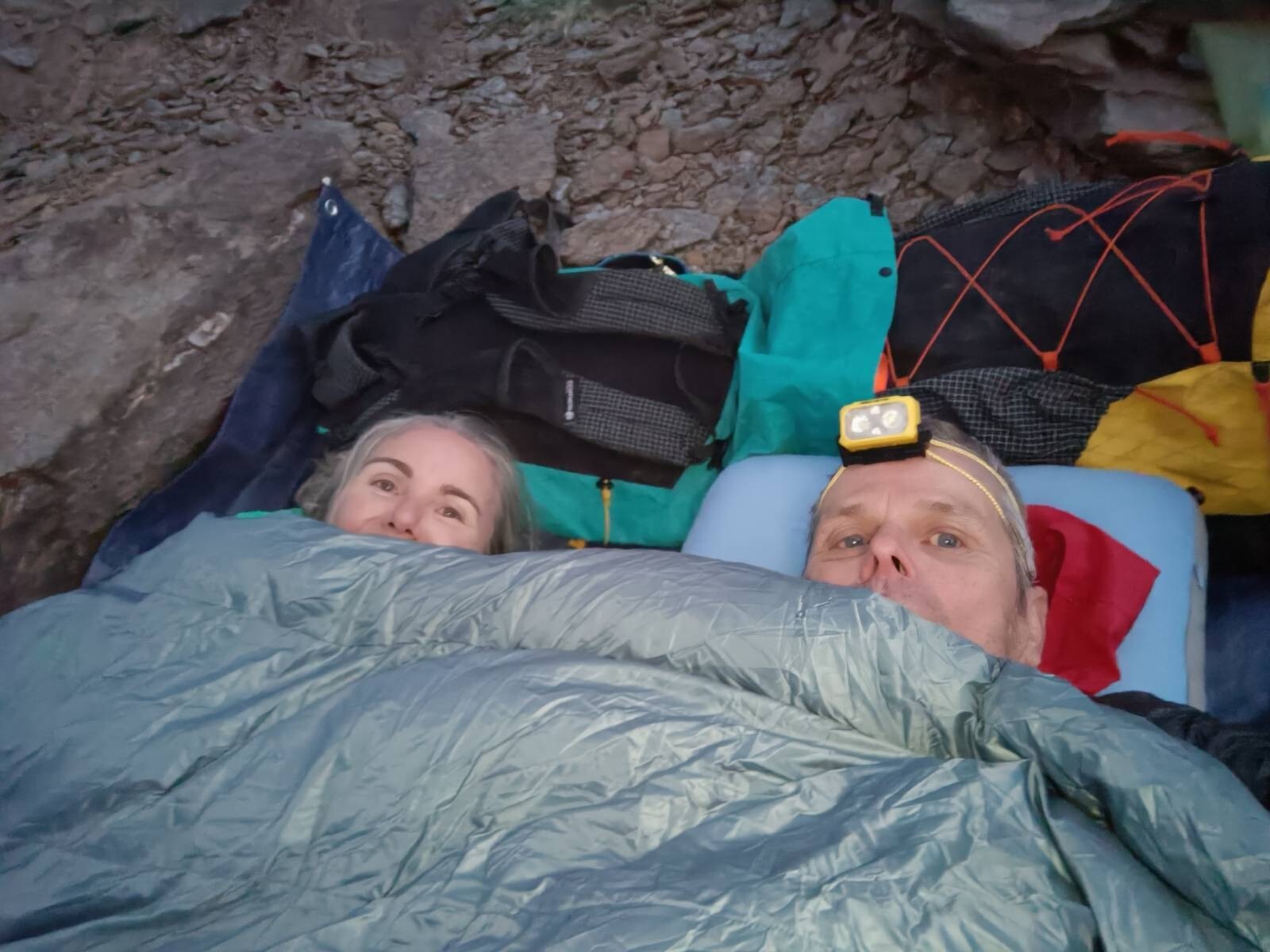 Two hikers wrapped in a sleeping quilt at a mountain bivouac site