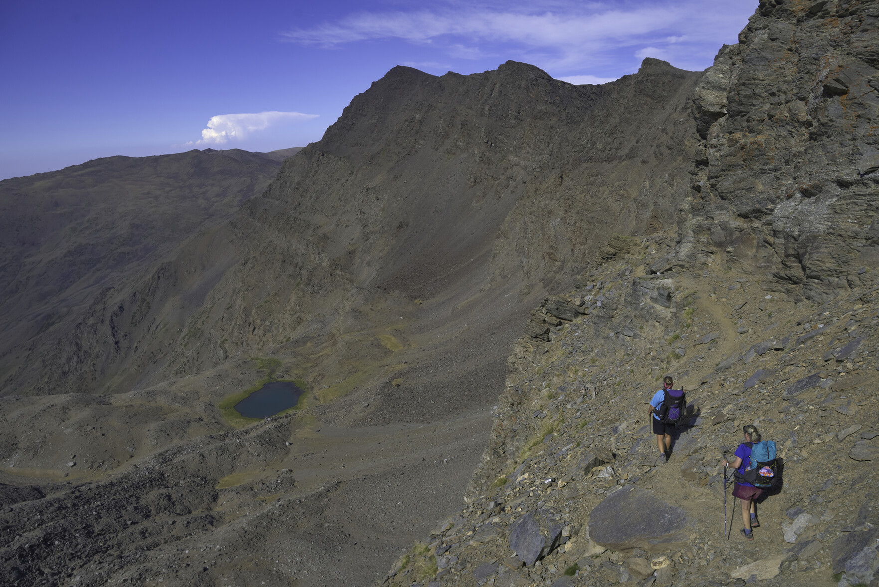 Some hikers walk along a narrow ledge across a mountain face. There is a blue lake far below