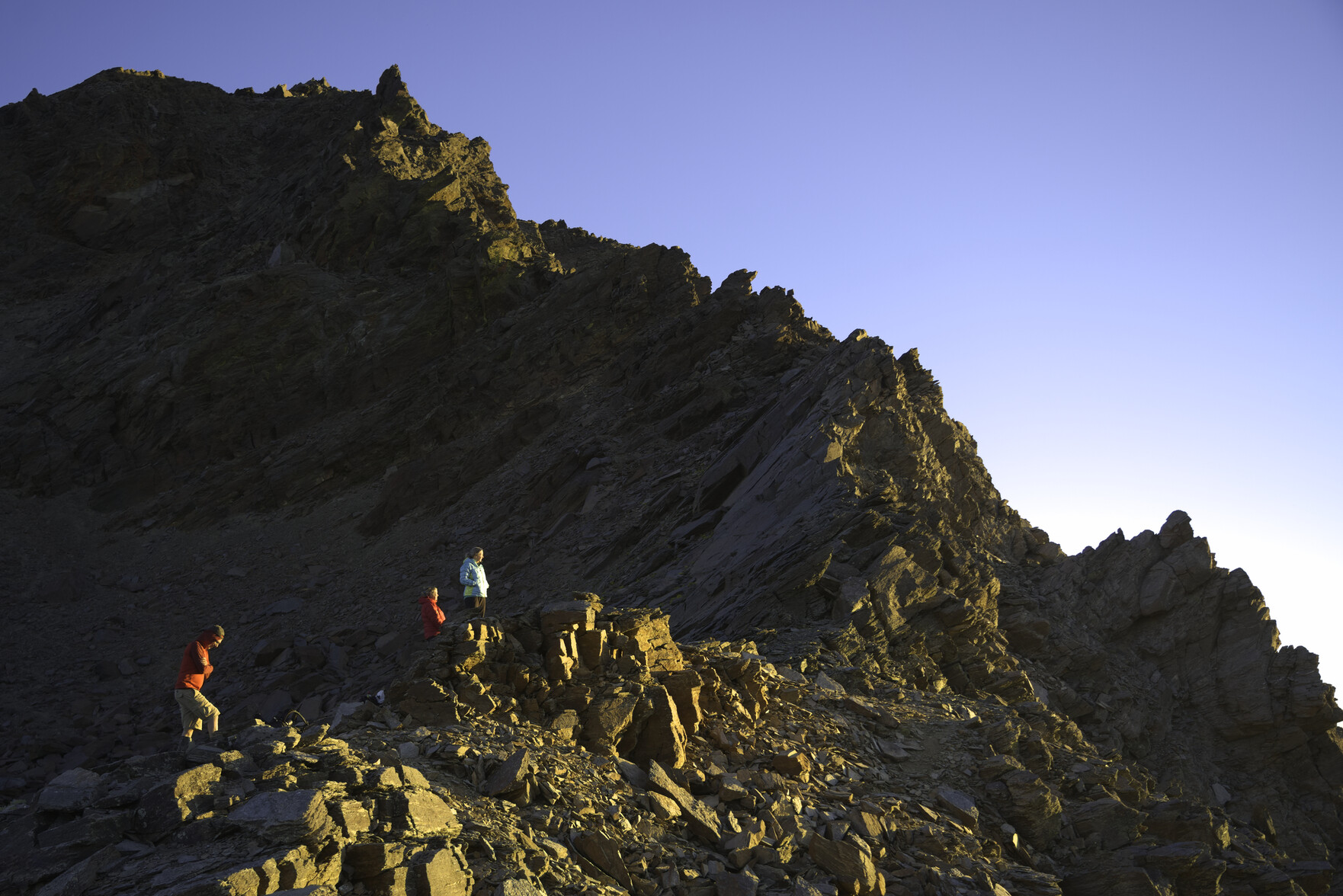 A group of people illuminated in the evening sunshine watch the sunset. behind rises a spiky mountain ridge