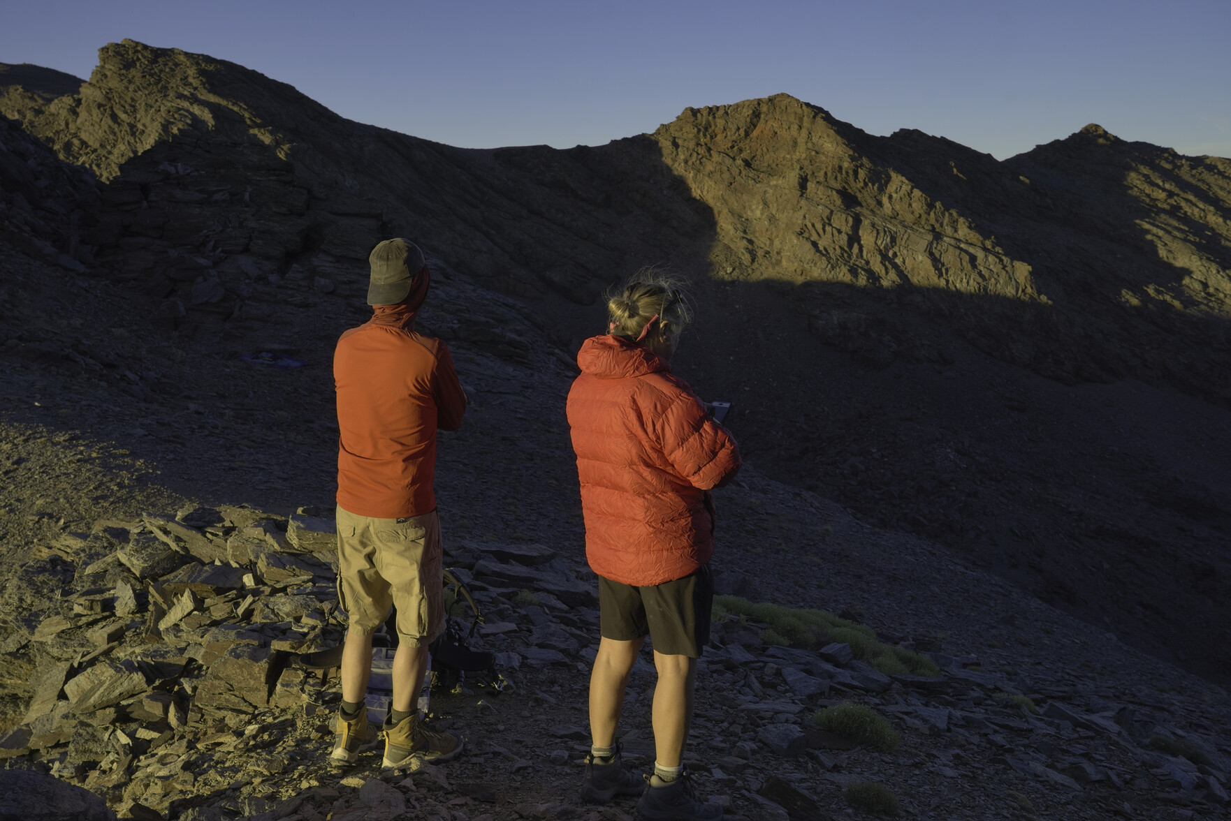 Evening sun shines on the back of two hikers and across to mountains in the background