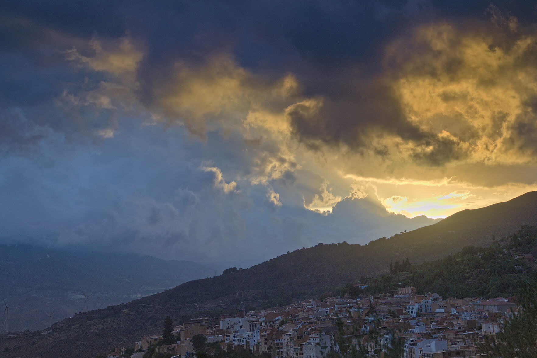 Dark storm clouds gather as the sun begins to set. Lower half of the image contains a spanish town at twilight