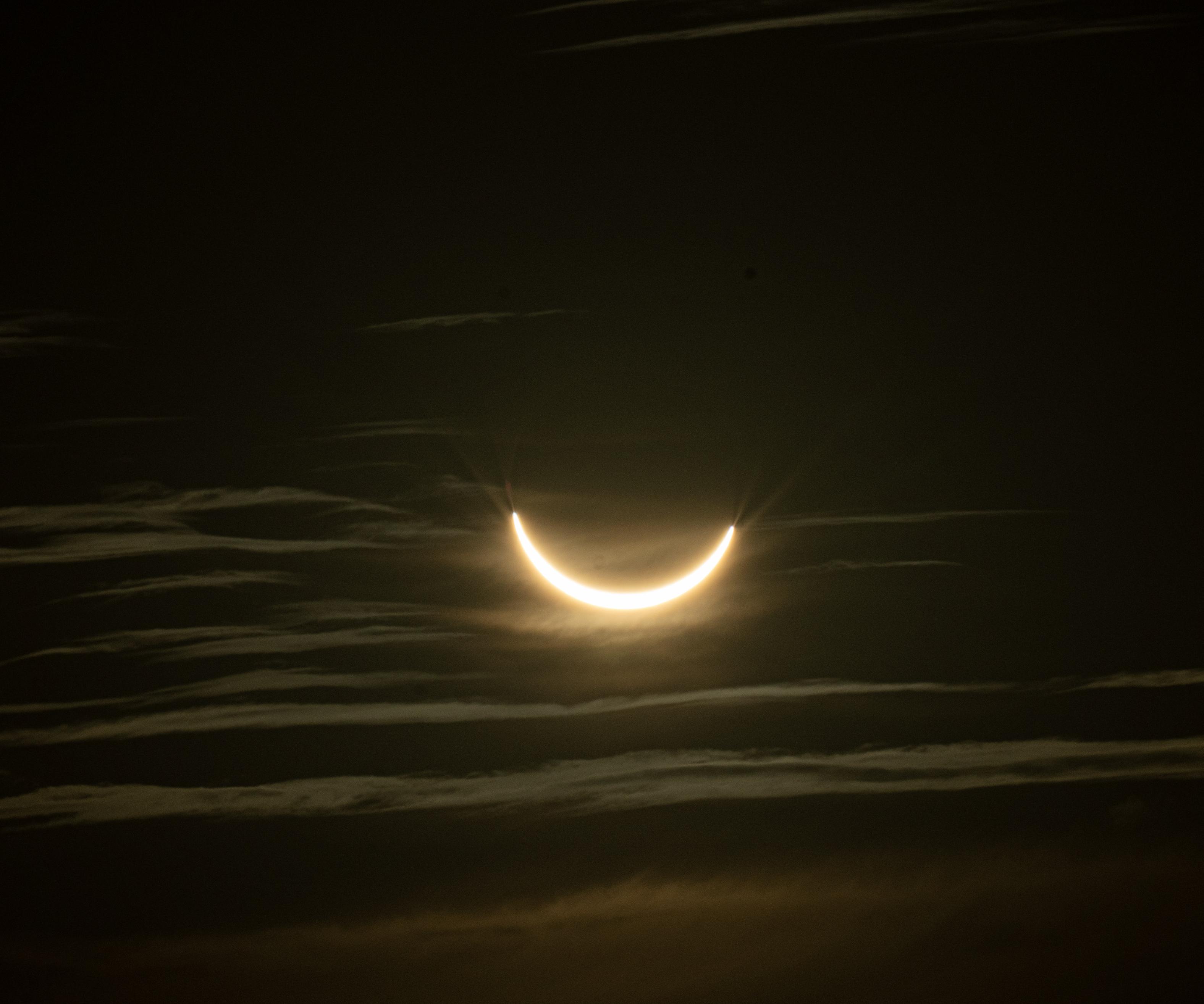 A partially covered sun during an eclipse. The lower half of the sun is uncovered and looks like a smile. You can almost make out the disc of the moon covering the sun. There are multiple rays coming out the edges of the smile. There are a few high light clouds just about shining through.